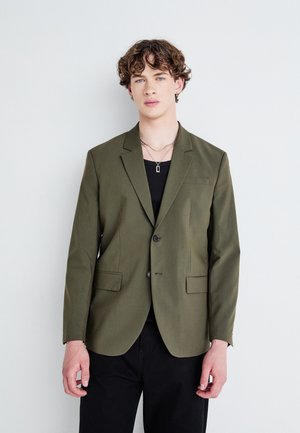 Mørk olivengrønn blazer med én knapp, klassiske lapeller og to frontlommer. Båret over en svart skjorte, kombinert med mørke bukser.