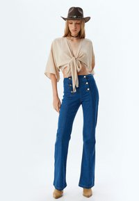 Top beige con lazo delantero y mangas anchas, combinado con pantalones de mezclilla azul de tiro alto y acampanados, con botones dorados y una textura suave. Sombrero marrón.