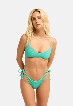 Blondynka o falowanych włosach, ubrana w turkusowy prążkowany bikini z wiązanymi bocznymi sznureczkami, stojąca na jednolitym jasnym tle.