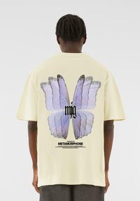 Yellow cotton t-shirt featuring a large blue butterfly graphic on the back, with black text below detailing the theme "METAMORPHOSE."