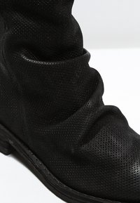 Bota de cuero negro perforado con textura arrugada y suela gruesa sobre un fondo blanco.
