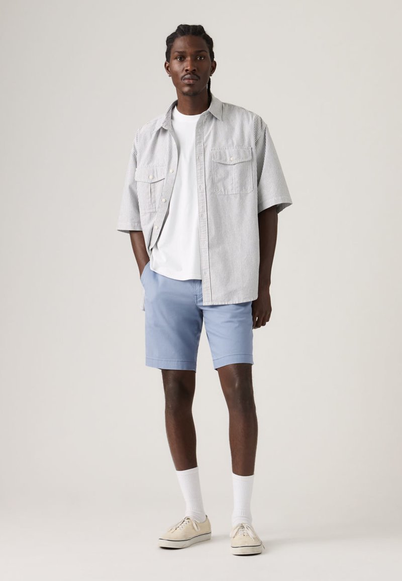 Homme debout les mains dans les poches, portant un T-shirt blanc, un short bleu clair, une chemise à manches courtes rayée, des chaussettes blanches et des baskets beige.