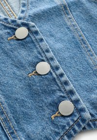 Primo piano di tessuto denim blu con tre bottoni metallici e cuciture beige su una cucitura del capo.