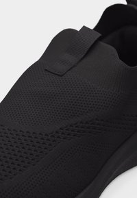 Μαύρο αθλητικό παπούτσι slip-on με πλεκτό διχτυωτό επάνω μέρος και τράβηγμα στη γλώσσα, με παχύ μαύρο πάτο.