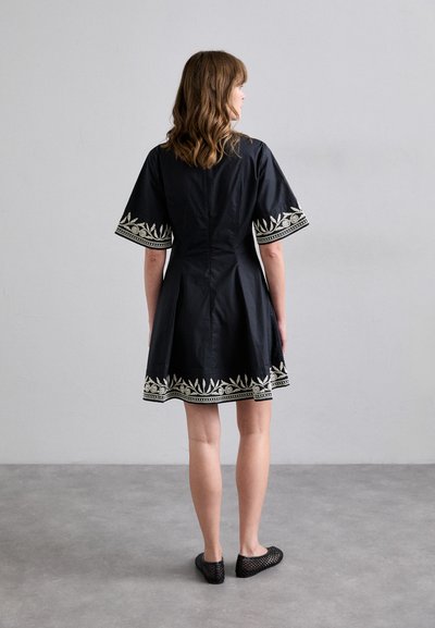 Vestido negro con mangas cortas, que presenta patrones de hojas bordadas en blanco a lo largo del dobladillo y los bordes de las mangas. Llevado con zapatos de malla negros.