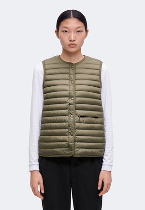 Jong persoon draagt een olijfgroene doorgestikte bodywarmer over een wit shirt met lange mouwen en zwarte broek, staand tegen een effen achtergrond.
