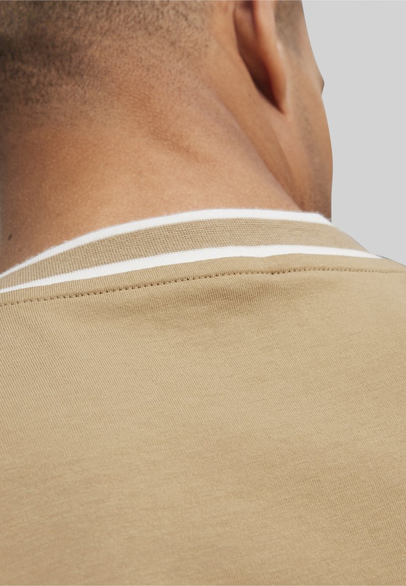 Camisa polo beige con un cuello acanalado con rayas blancas, textura suave y detalles cosidos en el escote, vista desde atrás.
