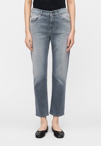 Femme portant un jean gris délavé droit, un haut noir et des ballerines noires, debout, les bras détendus le long du corps, sur fond blanc.