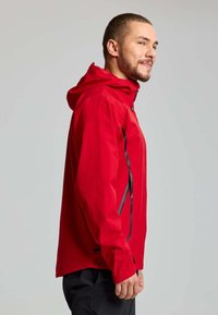 Veste imperméable rouge avec capuche, dotée de zips latéraux noirs et d'une coupe légèrement ample. Texture lisse et design léger.