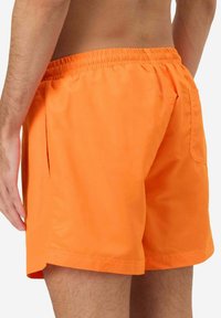Costumi da bagno arancioni vivaci realizzati in tessuto leggero, caratterizzati da una vita elastica e una tasca posteriore. Design corto e rilassato.
