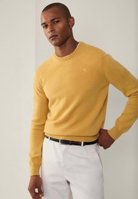 Žlutý svetr vyrobený z měkké pleteniny, kulatý výstřih, padnoucí silueta. Kombinovaný s kalhotami světlé barvy a tkaným tmavým opaskem.