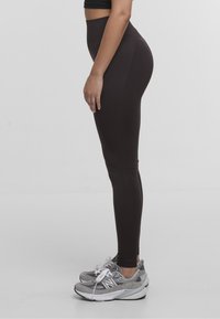 Leggings neri a vita alta realizzati in tessuto elasticizzato con una texture liscia, abbinati a scarpe sportive grigie con lacci bianchi e logo.