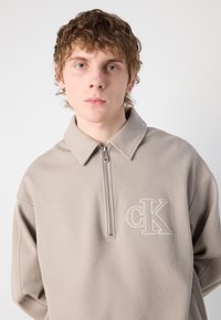 Pulover bej deschis cu fermoar la jumătate și textură în relief, având un guler ascuțit. Prezintă un logo brodat "CK" pe piept.