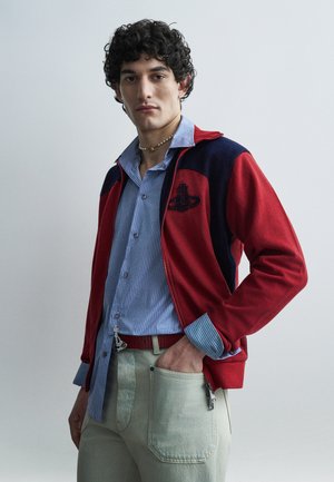 Homme aux cheveux bouclés portant une veste zippée rouge et marine par-dessus une chemise à rayures bleues et un pantalon clair, debout avec une main dans la poche.