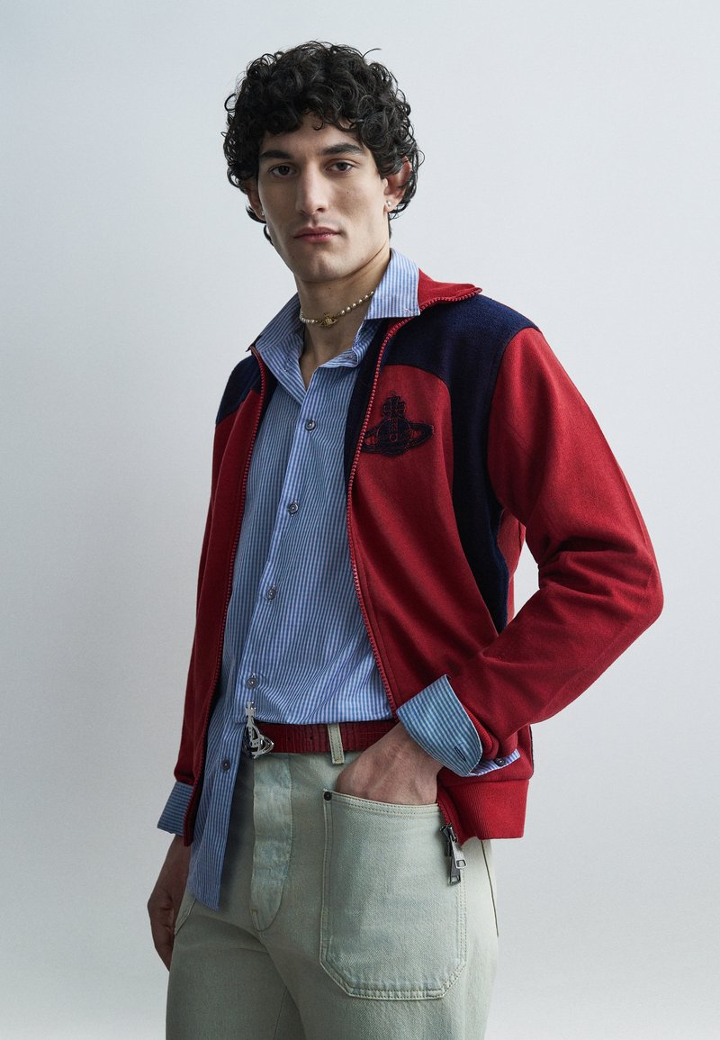 Άνδρας με σγουρά μαλλιά που φορά κόκκινο και μπλε μπουφάν με φερμουάρ πάνω από μπλε ριγέ πουκάμισο και ανοιχτόχρωμο παντελόνι, στέκεται με το ένα χέρι στην τσέπη.