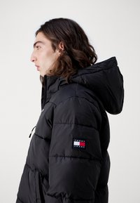 Sort pufferjakke med hætte, der har lynlås foran og vandret syninger. Et mærket patch er synligt på den venstre ærme.