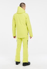 Veste et pantalon de ski jaune vif en matériau imperméable, dotés d'une capuche, de poches zippées et de poignets renforcés. Des bottes noires complètent l'ensemble.
