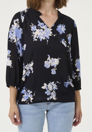 Kvinde iført sort V-hals bluse med blå og hvide blomstermønstre og lyseblå jeans, stående mod en ensfarvet baggrund.