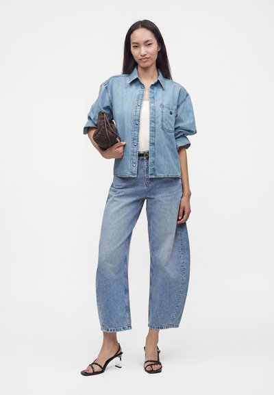 Jeune femme portant une veste en jean claire et un jean, tenant une pochette marron texturée, debout sur un fond blanc uni.