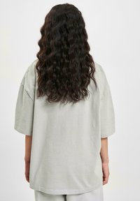 Oversized short-sleeve t-shirt in pale green cotton with a rounded hem and relaxed fit, viewed from the back. Curly hair visible.