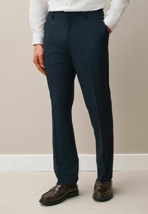 Mann trägt marineblaue formelle Hose, weißes Hemd und dunkelbraune Lederloafers, steht mit einer Hand in der Tasche.