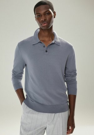 Camisa polo de manga larga gris con cuello, que presenta tres botones y puños acanalados, combinada con pantalones claros de rayas finas en gris.