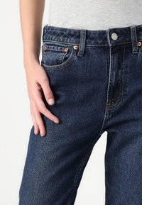 Dark blue denim jeans with a straight cut, five pockets, copper hardware, and contrasting orange stitching. Hand resting on the thigh area.
