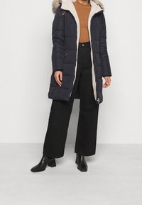 Lång marinblå puffjacka med pälsfodrad huva, med dragkedjestängning, sidofickor och kontrasterande beige insida. Bärs över brun stickad tröja och svarta byxor.