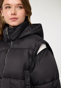 Piumino nero con collo alto, chiusura con cerniera e design senza maniche. Il tessuto ha una texture liscia e lucida con sezioni trapuntate.