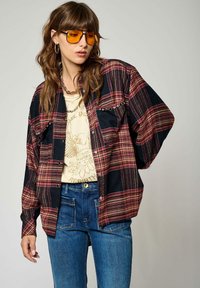 Chemise en flanelle à carreaux rouge et noir ; manches longues, à boutons, poches poitrine, associée à un t-shirt graphique beige et un jean bleu.