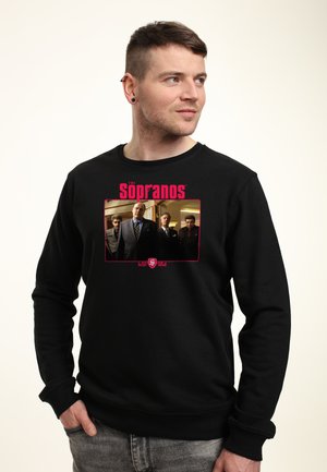 Mężczyzna w czarnej bluzie z czerwoną napisem "The Sopranos" i czterema mężczyznami w garniturach nadrukowanymi z przodu, patrzący w prawo.