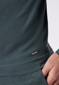 Dunkelgrüne Freizeitbluse aus strukturiertem Stoff, mit einem gerippten Halsausschnitt und einem kleinen silbernen Logo-Tag unten links.