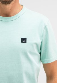 Light green short-sleeved t-shirt made from soft fabric; features a small black label with a white letter 'J' on the left chest area.