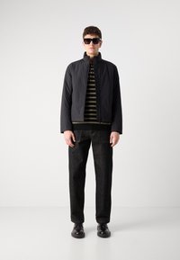 Jachetă neagră matlasată cu guler înalt peste o cămașă cu dungi, asortată cu pantaloni din denim închis și pantofi negri, purtată cu ochelari de soare mari.
