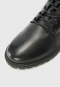 Primer plano de una bota de cuero negro con cordones y una suela texturizada, reposando sobre un fondo claro, enfatizando su superficie pulida.