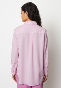 Blusa rosa chiaro con maniche lunghe, polsini abbottonati e dettaglio di yoke sul retro. Tessuto morbido e orlo leggermente curvo.