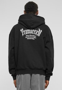 Svart hettegenser med snor i hetta, ribbestrikkede mansjetter og stor hvit grafisk tekst bak. Stoffet ser mykt og koselig ut.