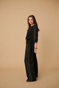 Femme debout de profil portant un pantalon noir à jambes larges et un haut assorti à rayures verticales beiges, bracelet manchette en bois au poignet droit.