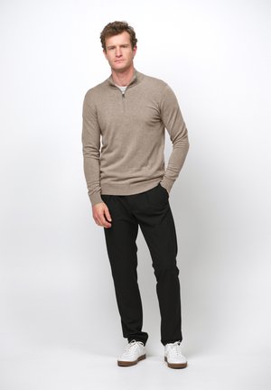 Mann steht mit den Händen in den Taschen, trägt einen beigen Pullover mit Viertelreißverschluss, schwarze Hose und weiße Sneaker vor weißem Hintergrund.