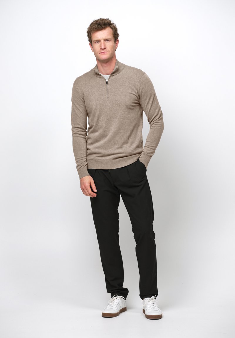 Mężczyzna stojący z rękami w kieszeniach, ubrany w beżowy sweter z krótkim zamkiem, czarne spodnie i białe trampki na białym tle.