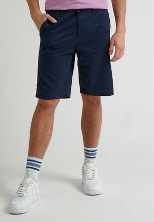 Mężczyzna w granatowych szortach, białych trampkach i białych skarpetkach z niebieskimi i szarymi paskami, stojący z ręką w kieszeni.