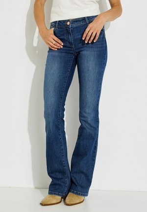 Persona que lleva vaqueros azules de corte bootcut, top blanco y botas de ante marrón claro con punta puntiaguda, de pie frente a una pared blanca lisa.