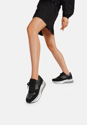 Scarpe sportive nere con pannelli in rete e camoscio. Suola intermedia spessa di colore bianco, punta rotonda e lacci neri. Movimento di camminata catturato con le gambe in movimento.