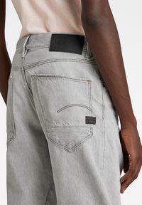 Jeans de denim gris claro con un corte relajado, que presentan un parche de logo de cuero negro, dos bolsillos traseros y detalles de costura sutiles.