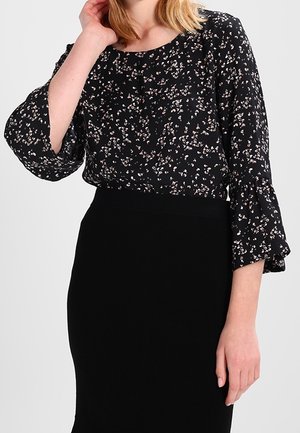 Vrouw die een zwarte blouse met bloemenprint draagt met 3/4 klokkende mouwen en een zwarte hooggetailleerde rok tegen een effen achtergrond.