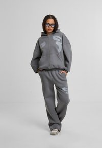 Sudadera con cremallera gris con grandes acentos gráficos, combinada con pantalones de chándal grises a juego. La tela parece suave y lisa, con un corte relajado.