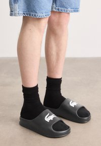 Sandale negre cu talpă plată, cu vârful deschis, având un logo alb de crocodil, purtate cu șosete negre până la glezne și pantaloni scurți din denim. Material din cauciuc neted.