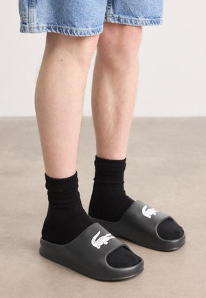 Des sandales noires à talon ouvert, arborant un logo crocodile blanc, portées avec des chaussettes de la même couleur et un short en jean. Matériau en caoutchouc lisse.