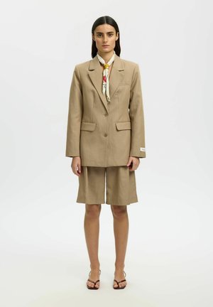 Femme aux longs cheveux bruns porte un blazer beige et un short assorti à longueur genou, accompagnés d'un foulard à motifs autour du cou et de chaussures ouvertes marron à talons.