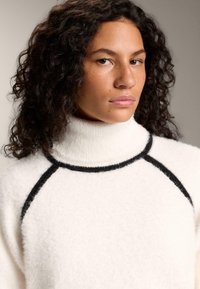 Maglione bianco a collo alto con bordi neri, realizzato in materiale morbido e peloso. Presenta una vestibilità comoda e maniche raglan.